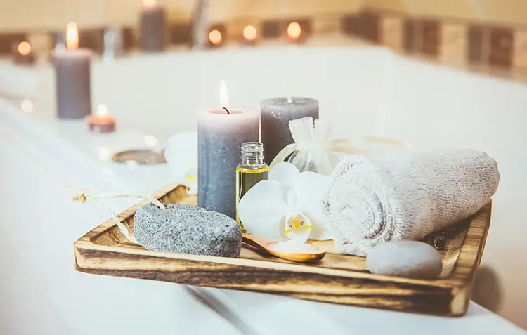 Spa tray with candles, hot towel, body scrubber, and oil on the edge of a bath tub